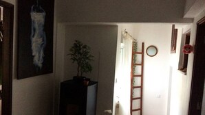 Ein schmaler Flur mit einer Topfpflanze, einer an der Wand angelehnten Leiter, einem Korbwesen und einem Bild an der Wand.
