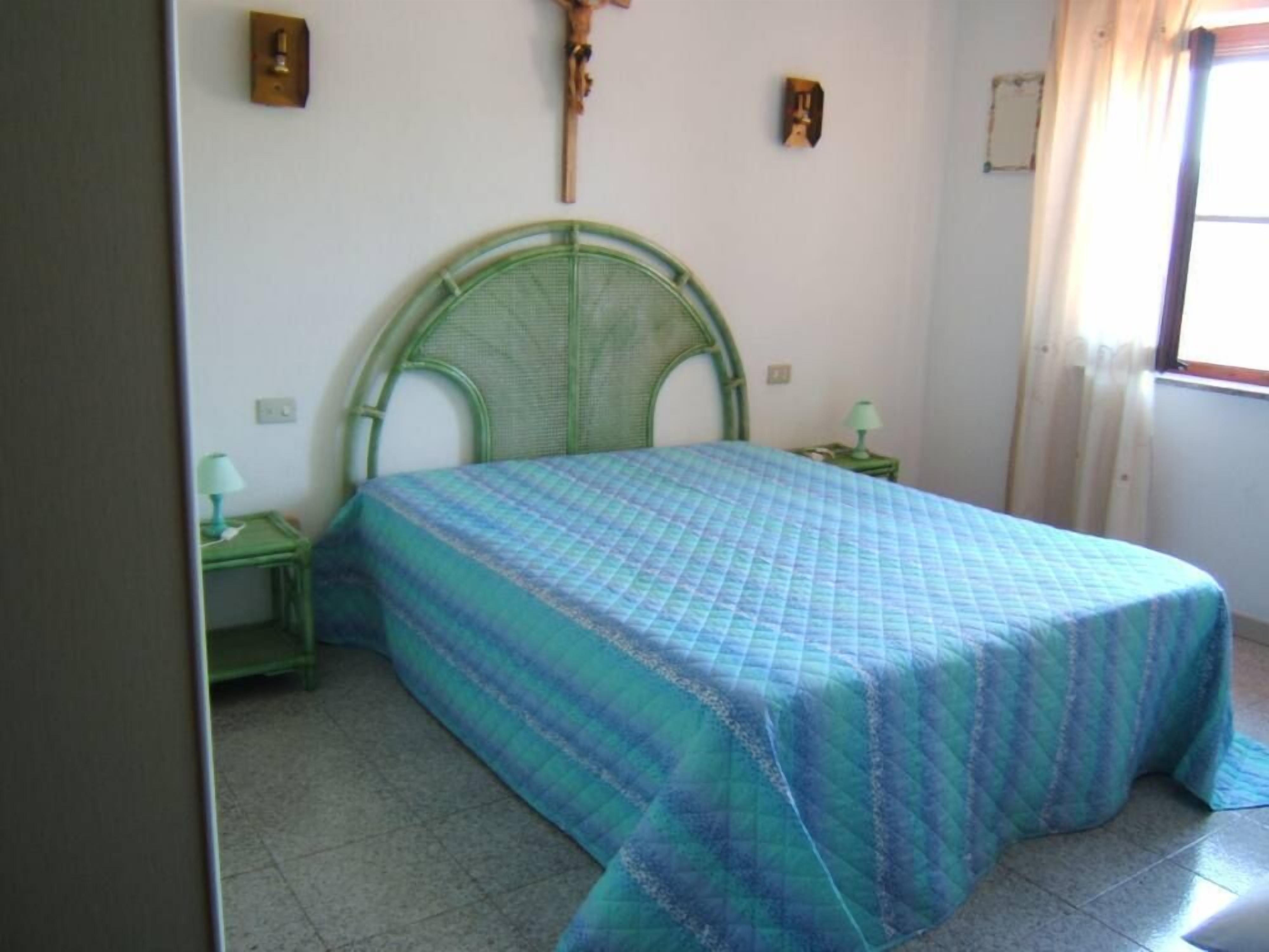 Une chambre à coucher avec une tête de lit en métal verte, un lit recouvert d’une courtepointe bleue, deux tables de chevet avec des lampes et une fenêtre avec des rideaux.