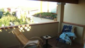 A balcony with a wicker chair and a small table, a second chair with a blanket, and a view of a garden and buildings. - Apartment/ flat - Budoni (Budoni)