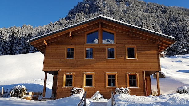 Un chalet en bois avec un toit à forte pente, de nombreuses fenêtres et une entrée recouverte de neige.