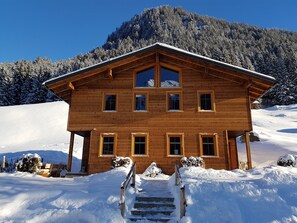 A wooden chalet with a steep roof, multiple windows, and a snow-covered entrance.