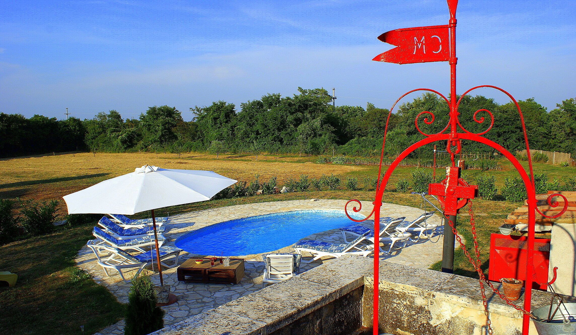 A swimming pool with lounge chairs and an umbrella, surrounded by a stone deck and a red metal fence.