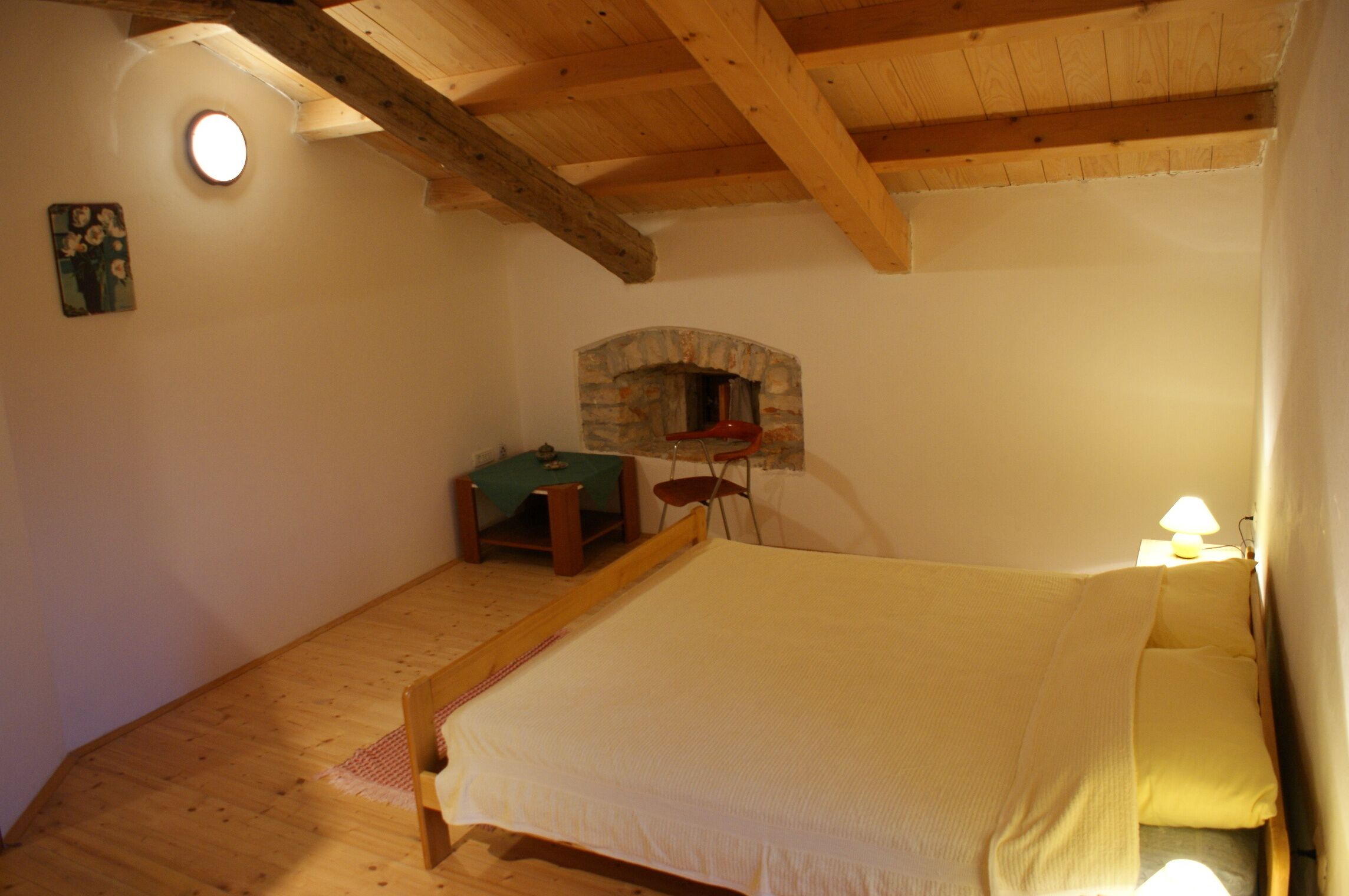 A bedroom with a bed, a stone fireplace, a small table with a chair, and a lamp.
