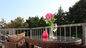Une table à plateau en verre, une chaise en osier, un vase rose et un verre à vin sont disposés sur un balcon doté d’une balustrade.