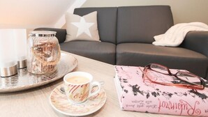 Un salon chaleureux avec un canapé gris foncé, un coussin blanc orné d’une étoile, un bocal en verre posé sur un plateau, une tasse de café et un livre rose sur lequel reposent des lunettes.
