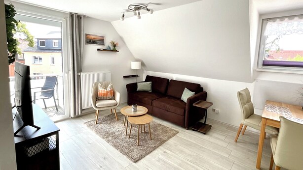 Un salon moderne avec un canap é marron, deux fauteuils, une table basse et un téléviseur. Il y a un balcon avec une chaise et une table, ainsi qu’une fenêtre donnant sur des immeubles.