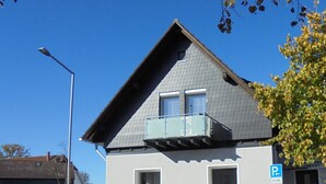 Une maison à deux étages avec un toit en pente, un balcon à l’étage supérieur et une petite fenêtre à l’étage inférieur.