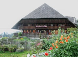 A traditional wooden building with a steeply pitched roof, surrounded by a wooden fence and blooming flowers. - Comfortable 3 bedroom apartment with rural charm in Emmental farmhouse (Trub)
