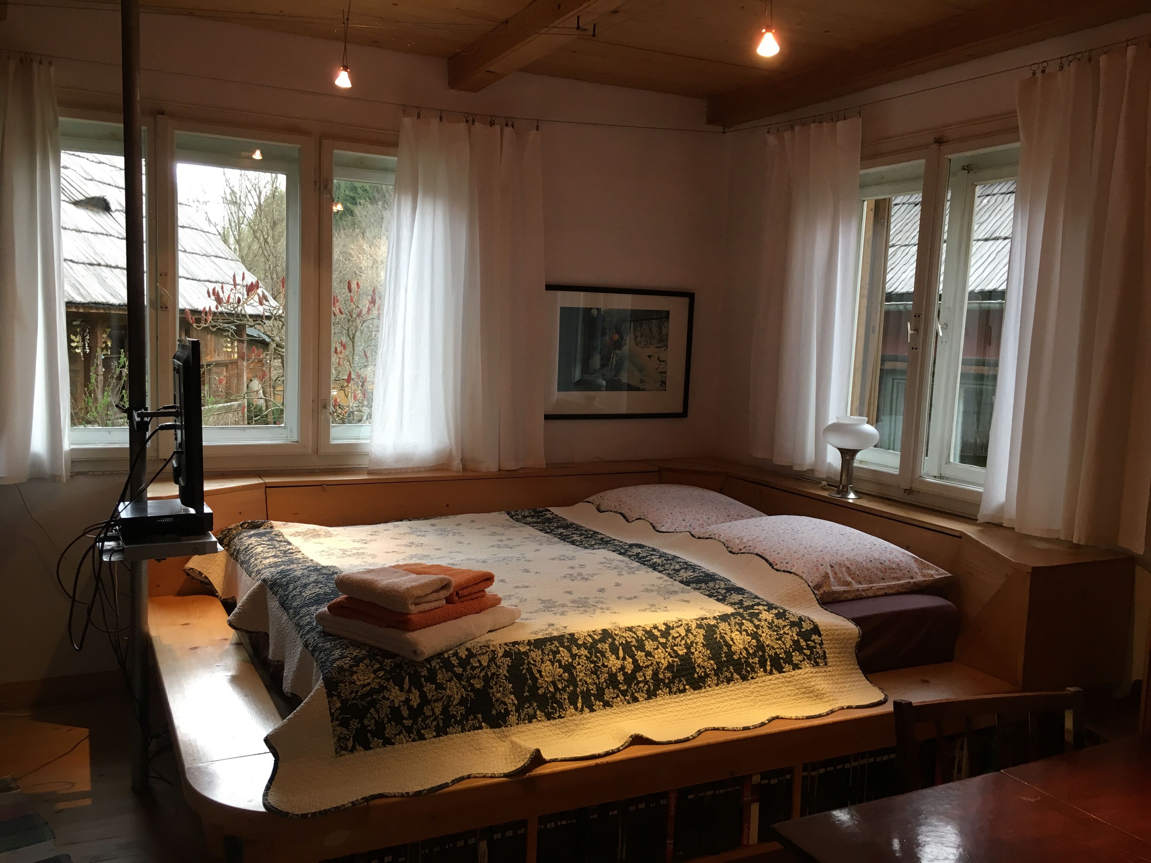 A bedroom with a large bed, a desk, and a chair. There are windows with curtains, a picture on the wall, and a TV on a stand.