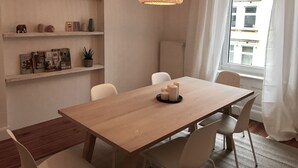 A dining area with a wooden table, white chairs, a hanging light fixture, and shelves with decorative items.