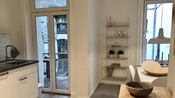 A modern kitchen with a sliding glass door leading to a balcony, a wooden dining table with a woven basket, and a white shelving unit with decorative items.
