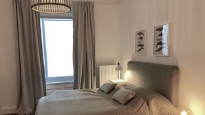 A bedroom with a large bed, a bedside table with a lamp, a window with curtains, and two wall-mounted pictures.