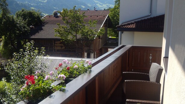 A balcony with wicker furniture, a table with a floral centerpiece, and a view of mountains and houses in the background. - Appartements Schweiberer, exclusive apartment Hambergblick (Ried im Zillertal)