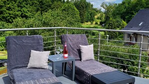 Uma varanda com duas poltronas, uma mesa pequena com uma garrafa e vista para uma paisagem verde.