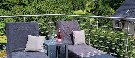 A balcony with two lounge chairs, a small table with a bottle, and a view of a green landscape.