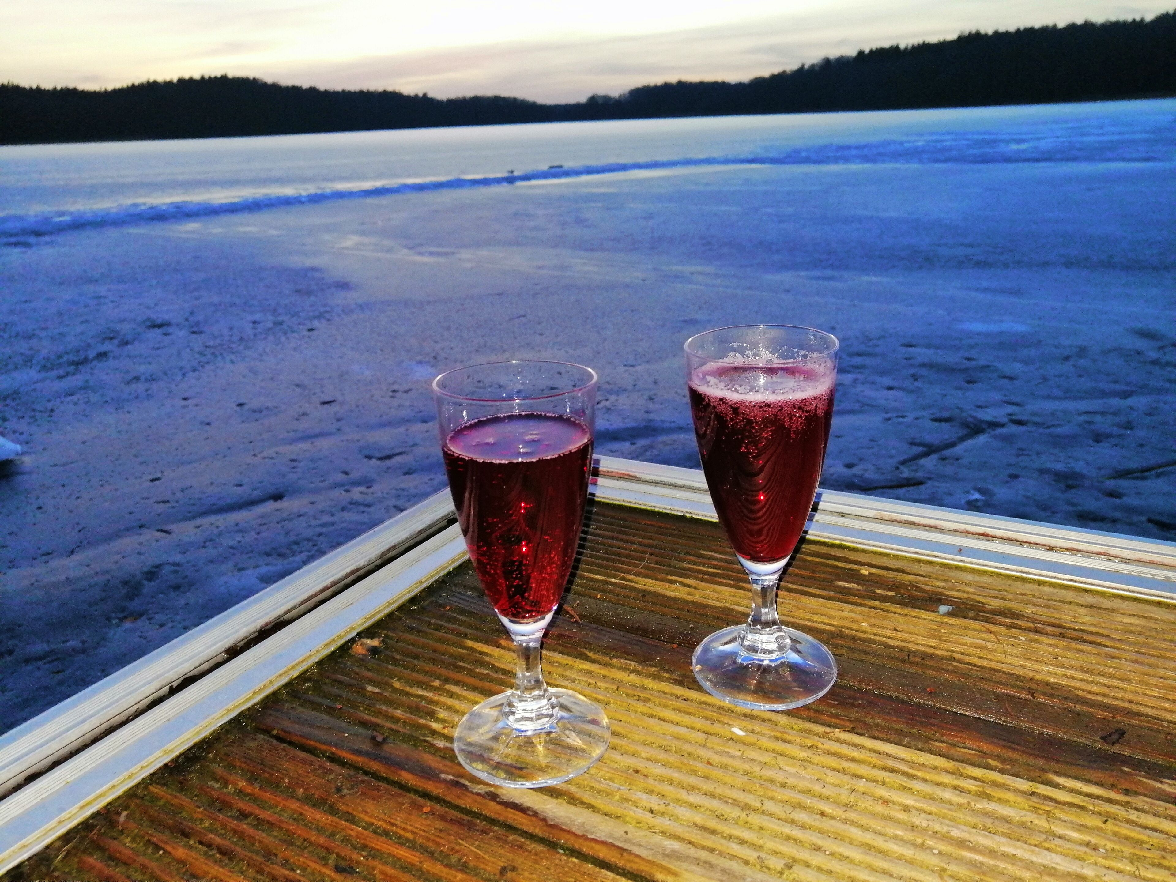 Två glas med röd dryck står på ett träbord med utsikt över en frusen sjö och en skog i bakgrunden.