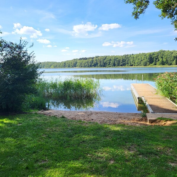 A lakeside view with a wooden dock extending into the water, surrounded by lush greenery and a forested hill in the background. - Holidays at Godendorfer See 300m, rowing boat, dog on request, fireplace, 2x toilet, 2 bedrooms (Godendorf)