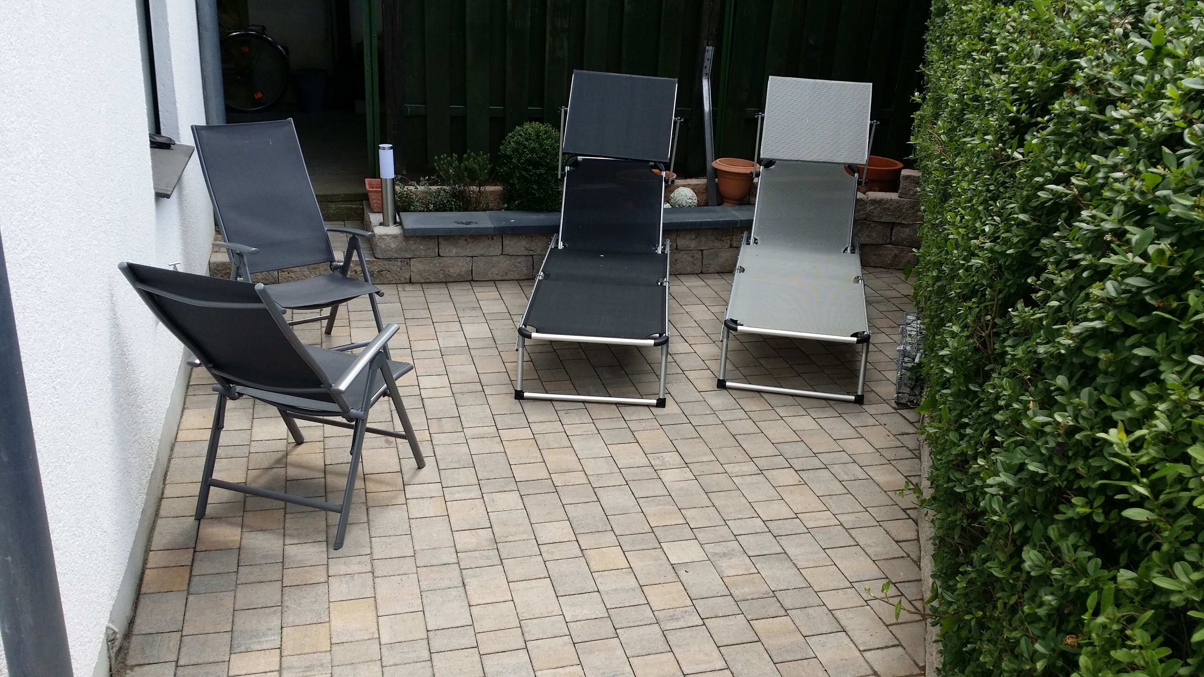 An outdoor patio with three reclining chairs and a table, surrounded by a hedge and a brick floor.