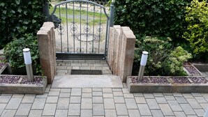 Une entrée fermée par un portail en métal et agrémentée de piliers décoratifs, entourée de jardins aménagés et d’un chemin pavé.