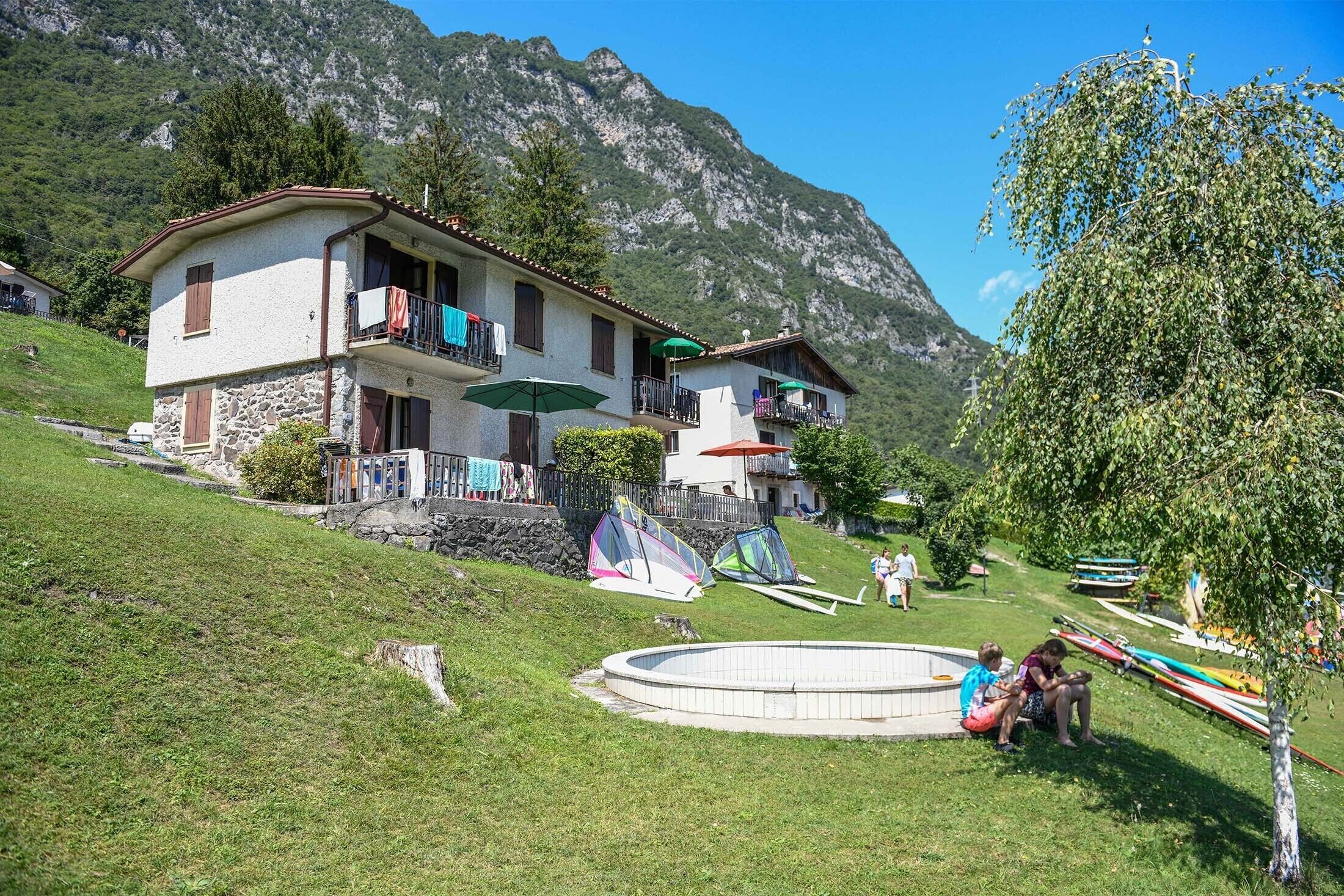 A two-story house with a stone facade, a balcony with a green umbrella, and a swimming pool in the yard.