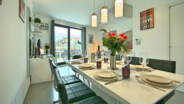 A modern dining area with a long table set for four, featuring a vase of red flowers, multiple wine glasses, and a view of the city through large windows. - PREMIUM VACATION HOME WITH SEAVIEW (Funtana)