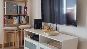 Un salon moderne avec un téléviseur à écran plat fixé au mur, une bibliothèque contenant divers livres et objets décoratifs, une machine à café et un petit coin repas avec deux tabourets.