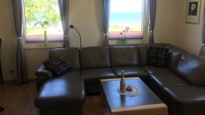 A living room with a brown leather sofa, a wooden coffee table, and a bottle of champagne. There are two windows with curtains, a floor lamp, and a framed picture on the wall. - Baltic Sea view from all windows, 50m to the beach (Zierow)