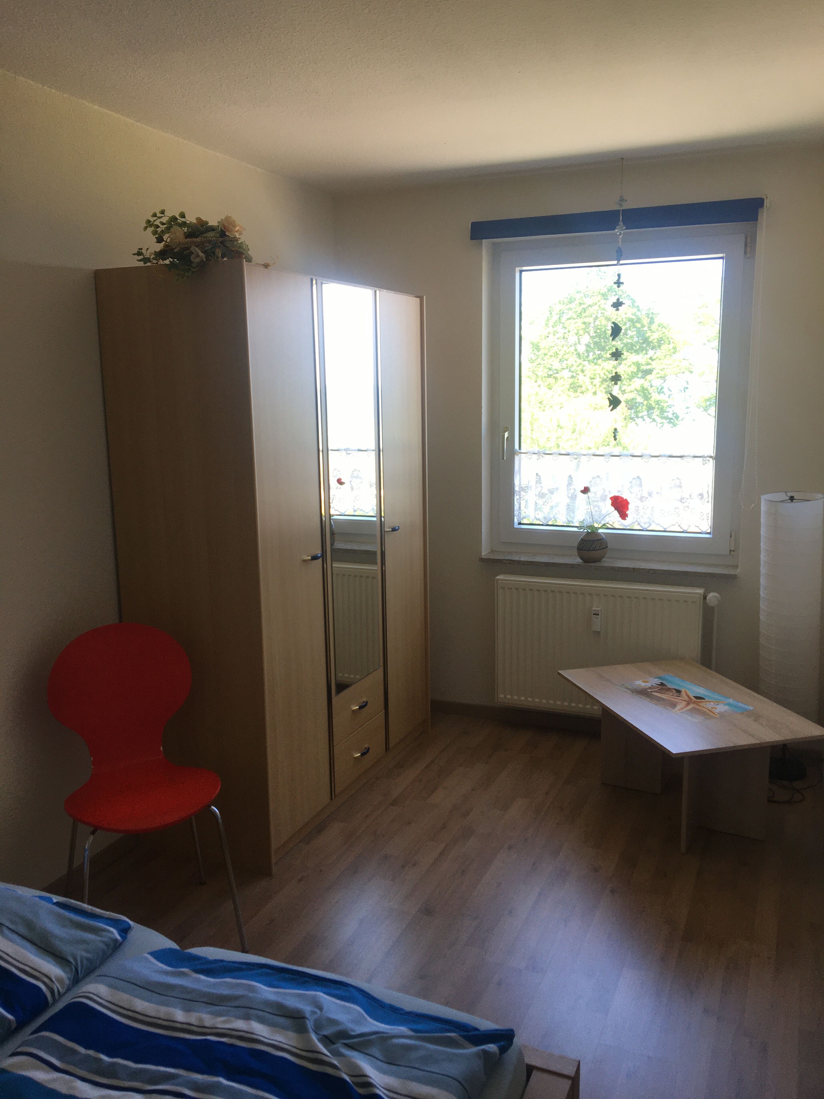 A room with a bed, a red chair, a wooden wardrobe, a small table, and a window with a view of trees.