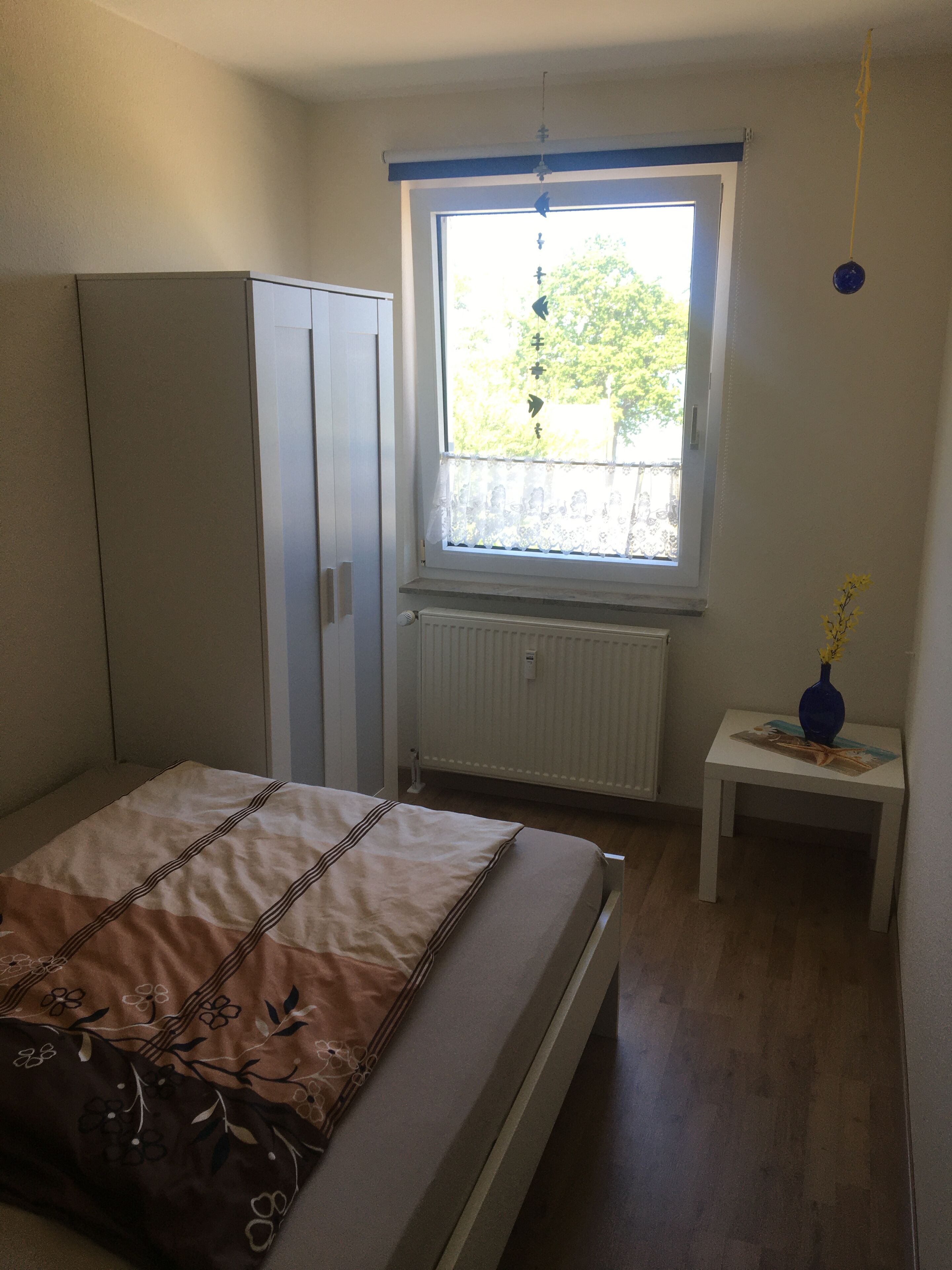Ein Schlafzimmer mit Bett, Kleiderschrank, Fenster mit Blick auf Bäume, Heizkörper und einem kleinen Tisch mit Vase.
