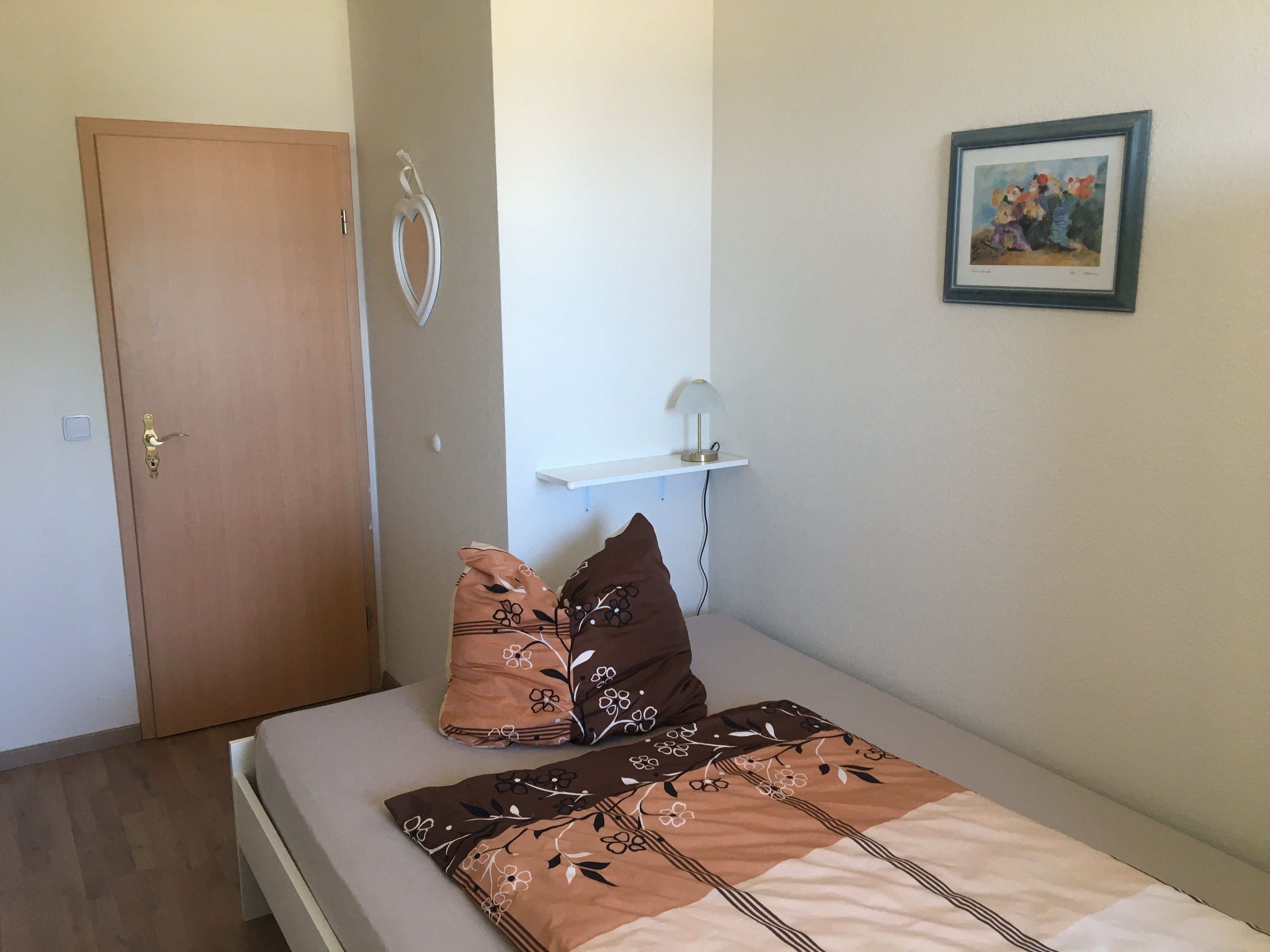 A bedroom with a bed, pillows, a door, a wall-mounted shelf, and a framed picture.