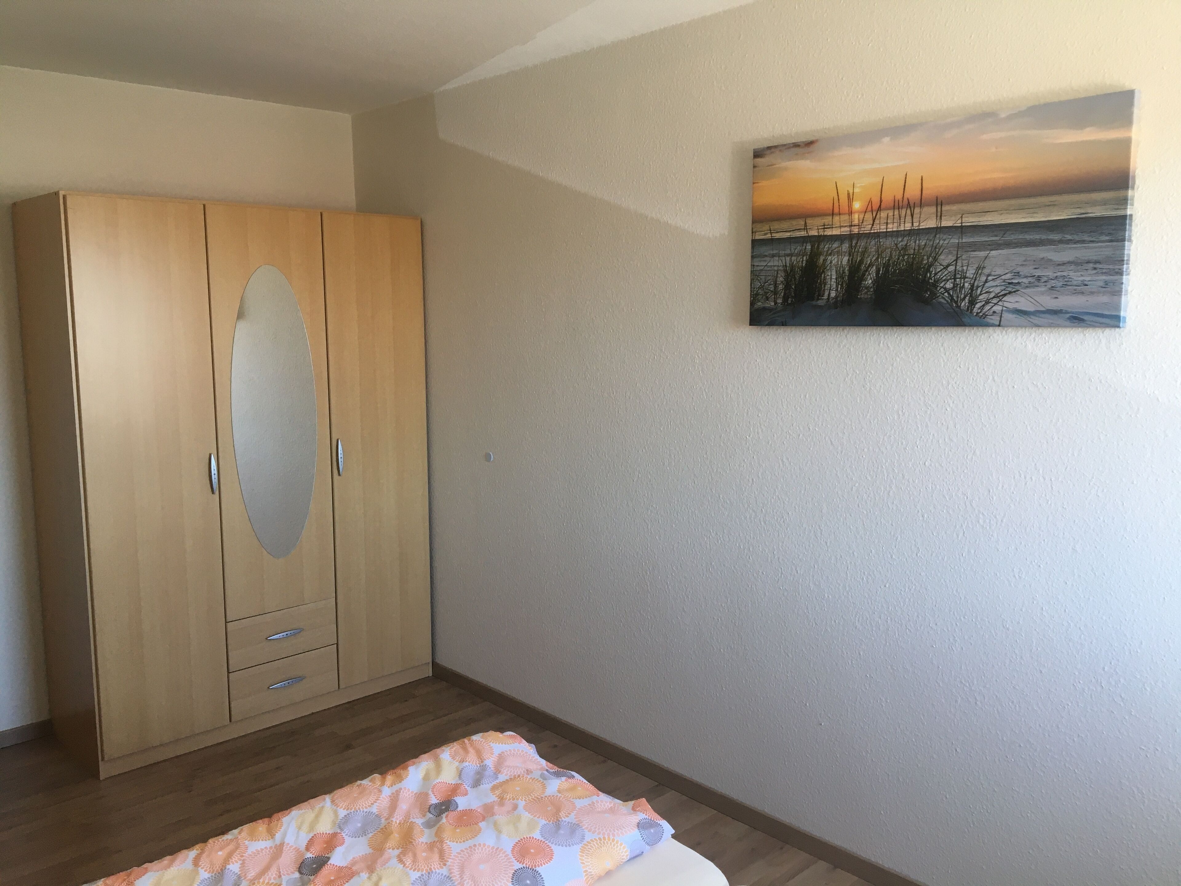 Une chambre avec une armoire en bois, un lit recouvert d’une couvre-lit à motifs et un tableau représentant un coucher de soleil accroché au mur.