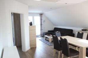 A modern living room with a dining area, featuring a wooden dining table, black chairs, a black sofa, and a large window with curtains.