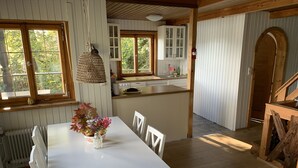 Uma área de refeições com uma mesa branca, cadeiras e um vaso com flores. Cozinha ao fundo, com armários e uma janela.