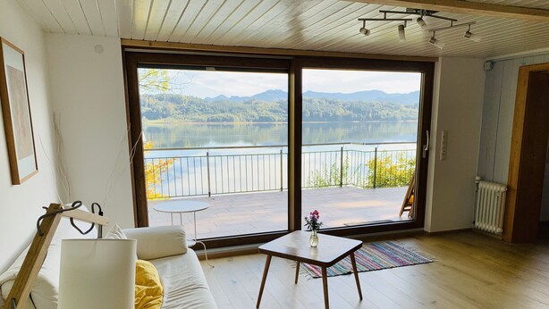 Quarto com sofá, uma pequena mesa e vista para um lago e montanhas através de grandes portas de correr.