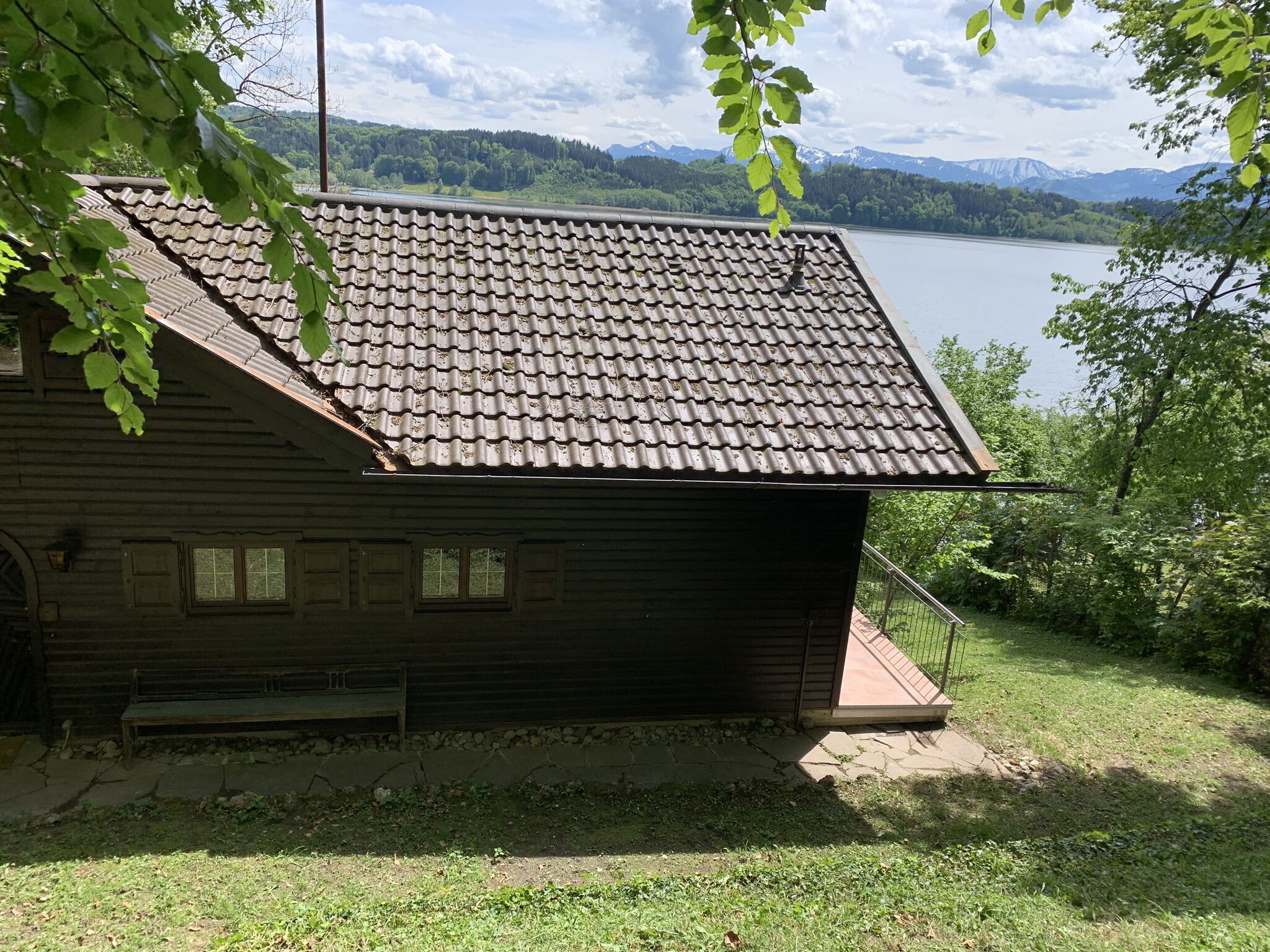 Una cabaña de madera rústica con techo de tejas, un banco de piedra y vistas a un lago y montañas.