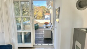 A room with a sliding glass door leading to an outdoor balcony with a table and chairs, a view of trees and a building, and a blue sofa inside. - Comfortable penthouse apartment with 5 stars, 50 m to beach (Mecklenburg Vorpommern)