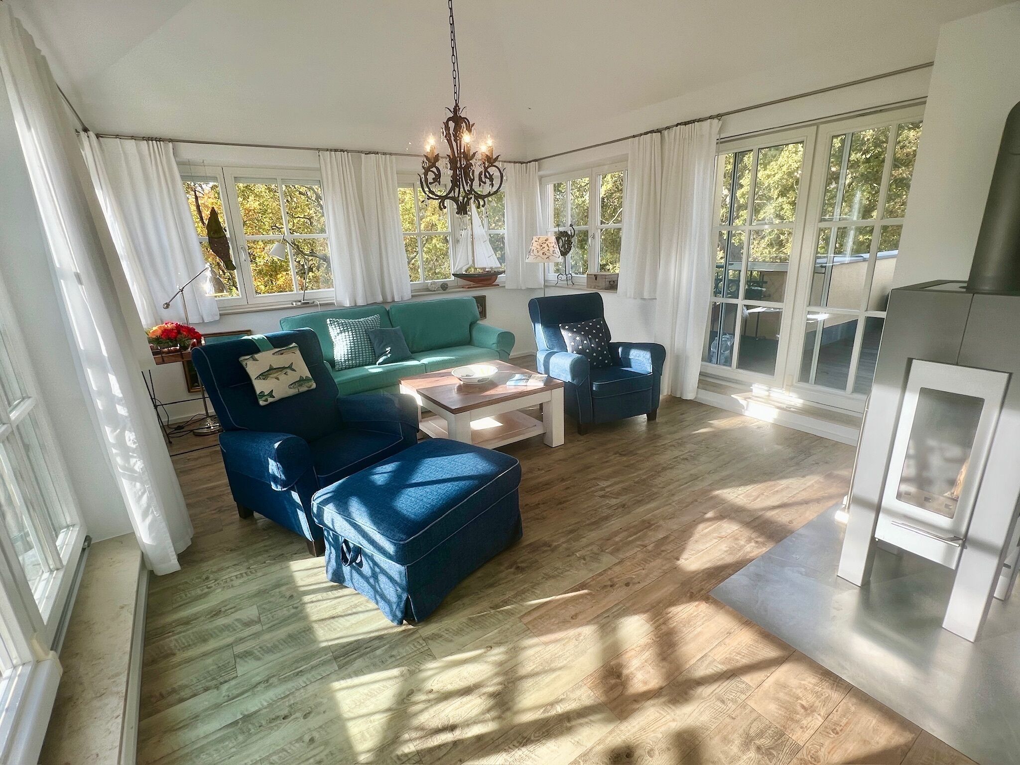 Amplia sala de estar con un sofá verde, sillones azules y una mesa de centro de madera. Ventanales grandes dejan entrar luz natural y ofrecen vistas al exterior.