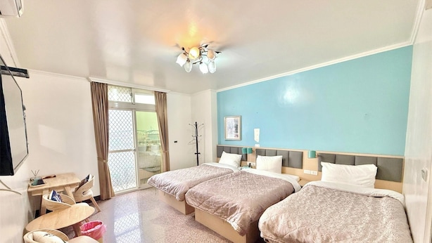 Habitación de hotel con dos camas, un escritorio y una silla. Se ve una ventana con cortinas, un ventilador de techo y una lámpara de pared.