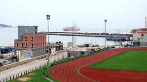Una pista de atletismo con superficie roja, una zona de césped y una masa de agua con un barco a lo lejos.