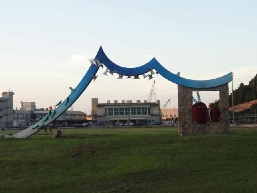 Un parque infantil con un tobogán azul y una estructura de escalada en un campo abierto, con edificios al fondo.