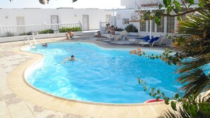 A swimming pool with a curved edge, surrounded by a tiled deck. There are lounge chairs and a few people in the pool. - Vv Apartamento Gran Vista Colony Club (Puerto del Carmen)