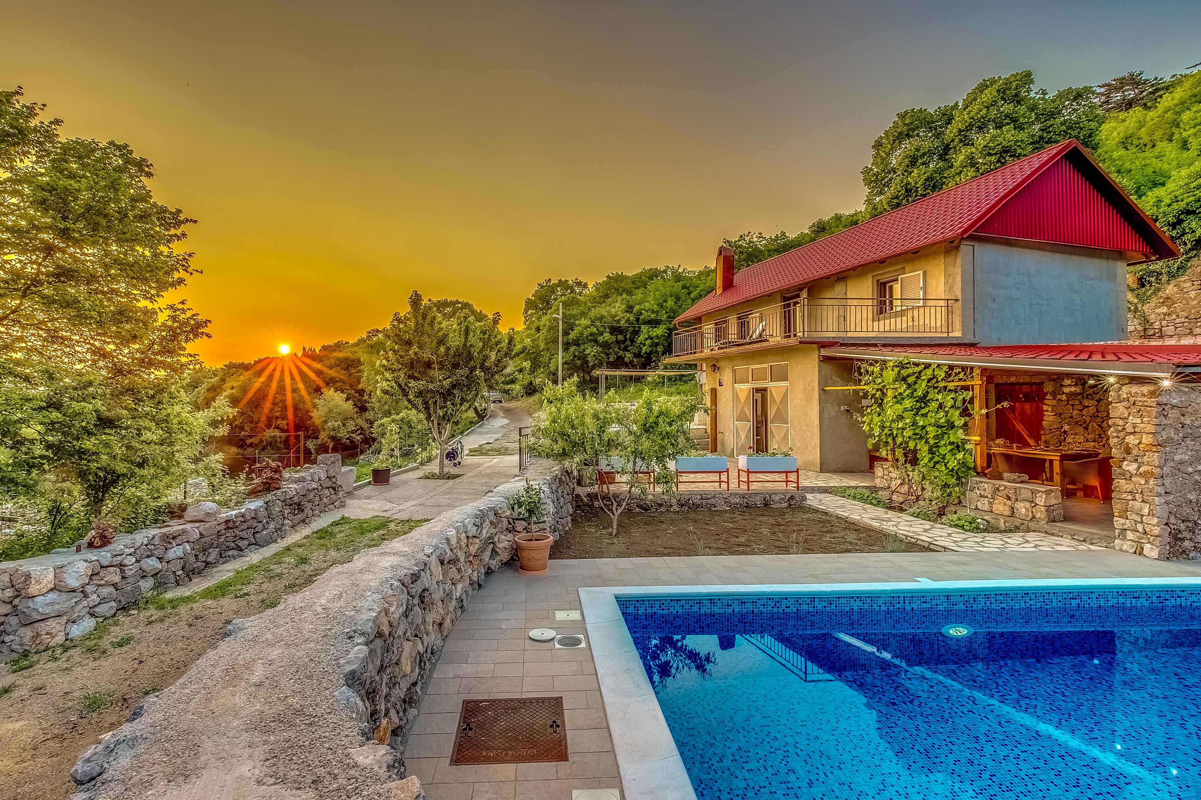 Une maison avec une piscine, entourée d’un mur de pierre et de verdure, avec un coucher de soleil en arrière-plan.