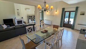 A modern living room with a glass dining table, white chairs, and a sofa. There is a fireplace, a television, and a bookshelf in the background. - Modern, large and beautiful villa with pool/garden in Azeitão south of Lisbon. (Azeitão, Setubal)