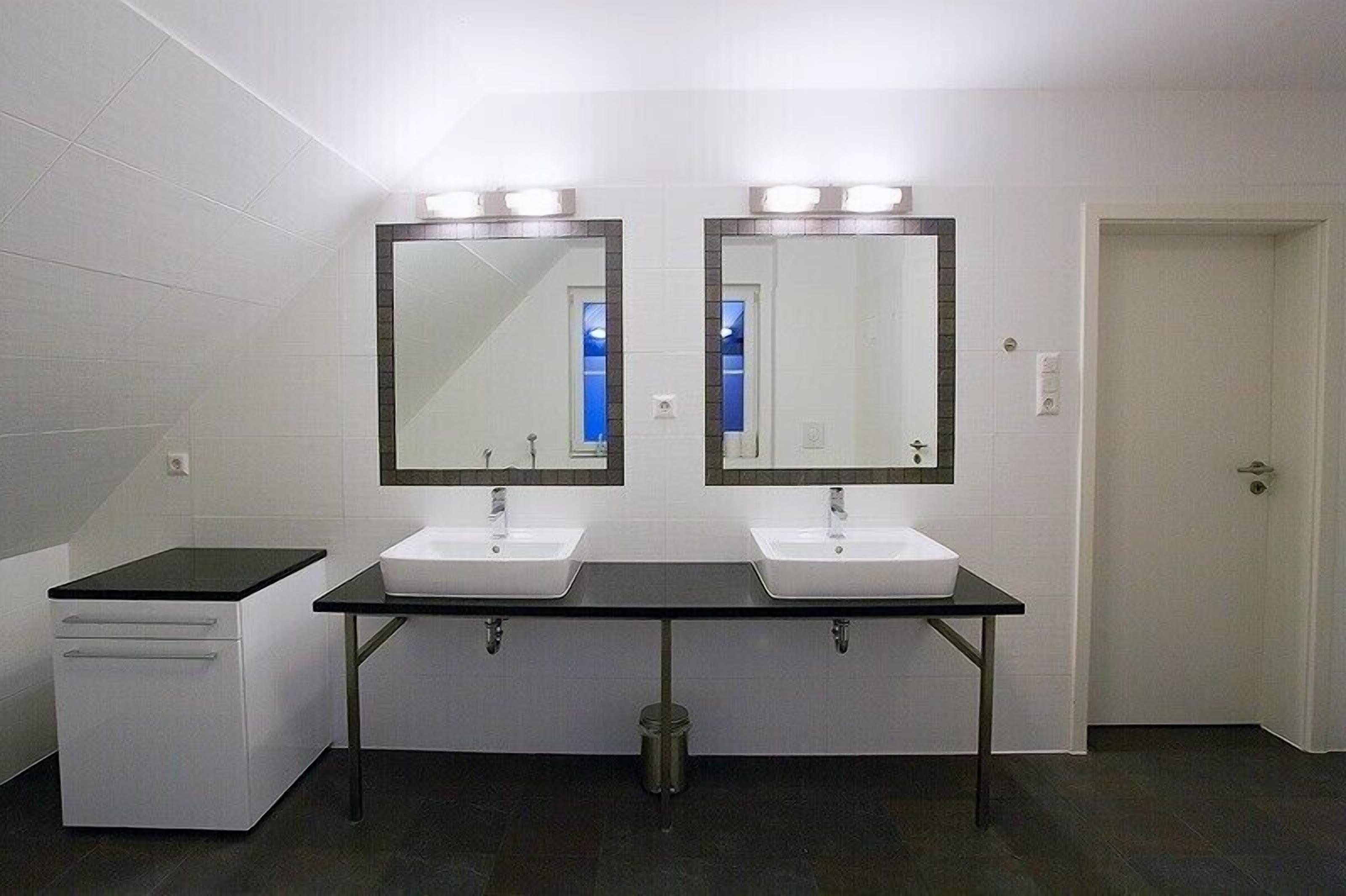 Een openbaar toilet met twee wastafels, een spiegel boven elke wastafel, en een prullenbak in het midden.