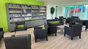 Un'area lounge moderna con divani e poltrone, una libreria con riviste e vista su uno spazio giochi esterno.