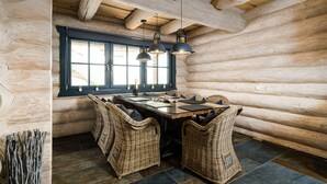 Interior de una cabaña de troncos con una mesa de comedor de madera, sillas de mimbre y lámparas colgantes.