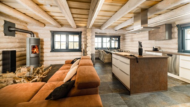 Interior de una cabaña de troncos con sofá, chimenea, cocina y ventanas amplias.