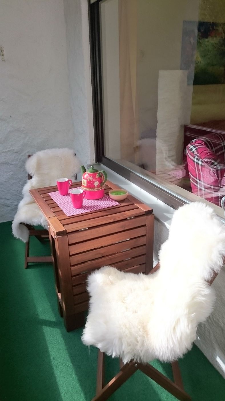 Une petite table en bois, sur laquelle on a posé un set de table rose, deux tasses roses et un objet décoratif, le tout sur un balcon recouvert d’une moquette verte.
