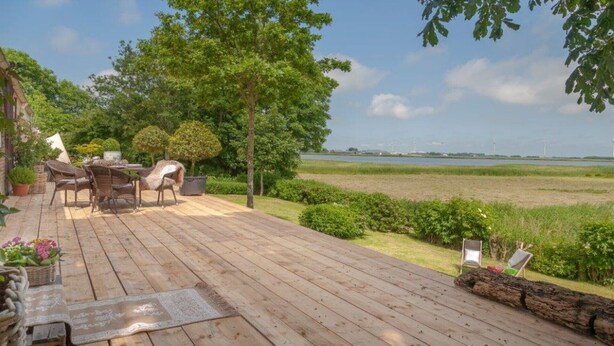 En træterrasse med udendørs møbler, omgivet af grønt og udsigt over et vandareal med vindmøller i det fjerne.