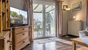 Ein Zimmer mit einem Holzschrank, einem Fernseher, einem Tisch und Blick auf eine Terrasse mit Stühlen und einem Tisch.